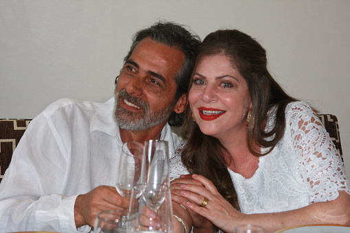Married couple happily toast with champagne, wearing white. She looks at the camera, he looks away, both smiling