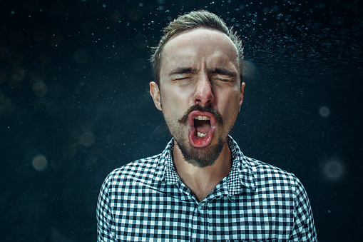 Young funny handsome man with beard and mustache sneezing with spray and small drops, studio portrait on black background. Comic, caricature, humor. illness, infection, ache. Health concept