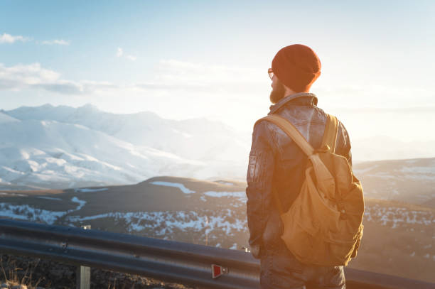 istock Bearded tourist hipster man in sunglasses with a backpack stand back on a roadside bump and watching the sunset against the background of a snow capped mountain 992204228