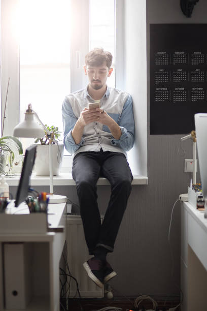 istock Young stylish man sitting on the window with phone in hands 992312026