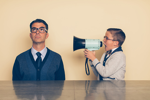 A young nerd boy in eyeglasses and a bow tie is yelling at his dad through a megaphone. His dad is looking up at the ceiling and ignoring what the son is saying. The son is frustrated with his dad because he won't listen as there is a generation gap.