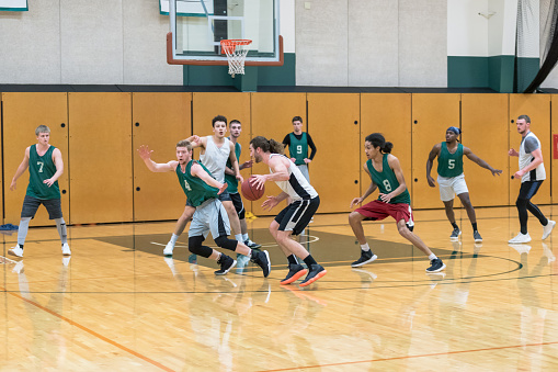A men's college basketball team practices in the gym. They are scrimmaging and a player in the foreground is driving to the basket while the defense tries to stop him and his teammates try to get open.