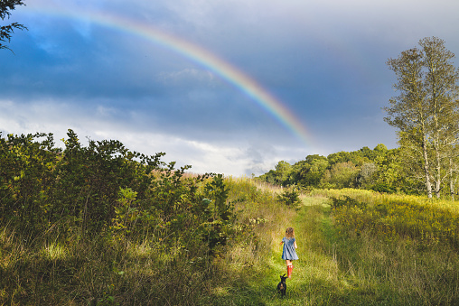 Over the rainbow... Adorable little girl in a blue dress and red boots in a vast nature field meadow area with a stunning rainbow in the sky. She is headed for adventure or maybe to find home?