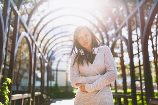 portrait of beautiful young woman wearing light casual clothes. girl standing under sunshine archway with headphones on her neck in city park in street outdoors on spring nature. lifestyle concept. - child office chaos computer monitor imagens e fotografias de stock