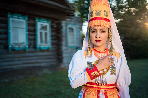Cute young woman dressed in women's traditional clothes and jewelry which are recreated according to medieval samples of the Mari people. The young woman stands on a lawn before the wooden country cottages. She is calmly looking at the camera. Shooting in a countryside on a summertime at a sunset, Republic of Mari El in Russia's Volga region