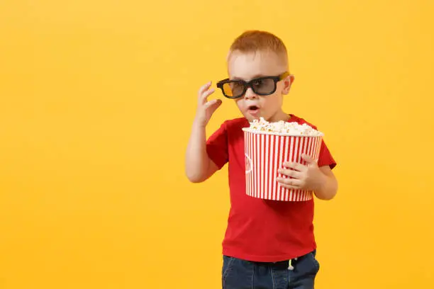 Little cute kid baby boy 3-4 years old in red t-shirt, 3d imax cinema glasses holding bucket for popcorn, eating fast food isolated on yellow background. Kids childhood lifestyle concept. Copy space
