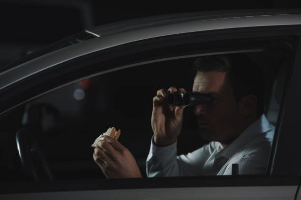 istock male private detective doing surveillance by binoculars and eating sandwich in car 992786532