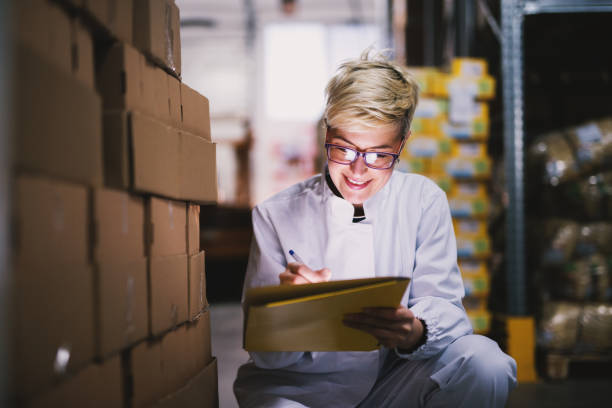 istock Young beautiful joyful female worker is filling info on the tablet she is holding while kneeling beside stacks of boxes. 992810414