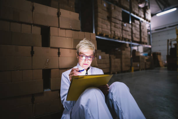 istock Young beautiful female worker is sitting leaned on a stack of boxes in big storage hangar while checking her notes on tabled on yellow folder leaned on her knee. 992810450