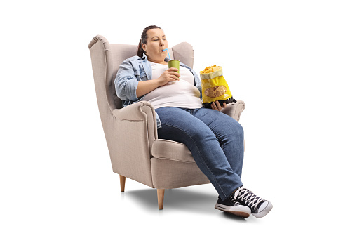 Overweight woman with a drink and a bag of chips sitting in an armchair isolated on white background