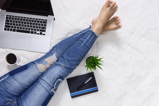 Woman wearing denim jeans sitting on bed and working with laptop. Domestic life lifestyle concept.