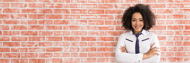 Smiling african businesswoman standing arms crossed in front of brick wall, looking at camera.