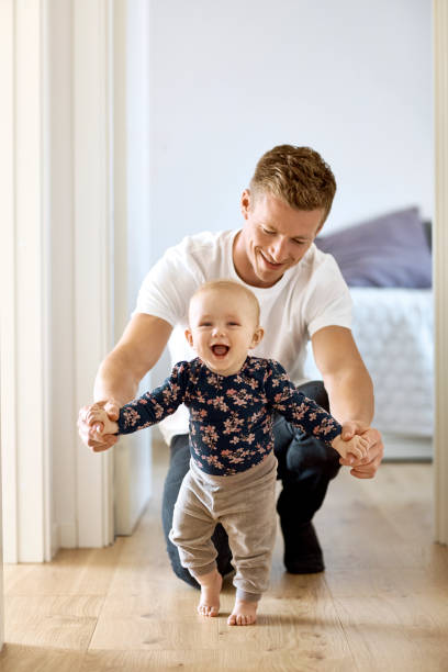 Father teaching son to walk in corridor at home Father teaching baby son to walk in corridor. Portrait of cheerful little boy holding hands of man at home. They are wearing casuals. first steps stock pictures, royalty-free photos & images