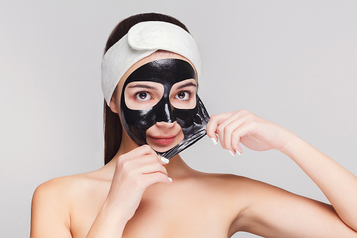 Skin care. Portrait of young girl peeling off charcoal mask from her face. Purifying spa treatment, white studio background, copy space