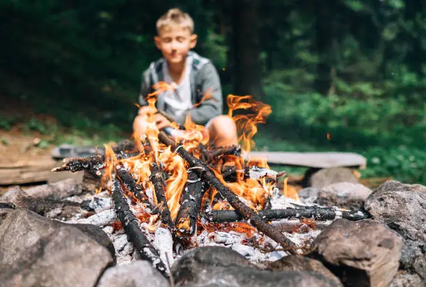 Photo of Boy sits near campfire. Summer camping time