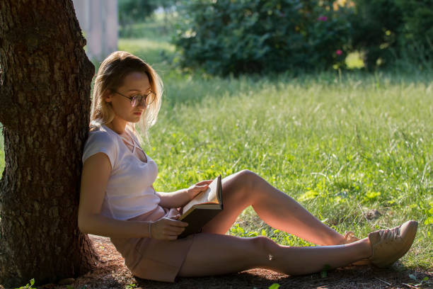 istock Young attractive woman reading book under the tree in park at su 992888990
