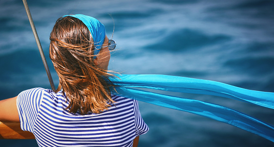 Closeup rear view of a mid 20's woman on a sailboat looking at the distance while her headscarf is flapping in the wind. Copy space on the right.