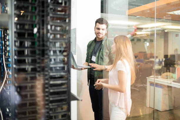 A man in a green jacket and a woman in a white shirt smile and discuss over a laptop in a modern office setting, with a server rack visible in the foreground.