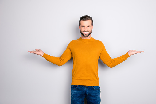 Portrait of delightful confident expert attractive bearded guy demonstrating balance  giving two options wearing mustard color sweater jeans isolated on gray background copyspace