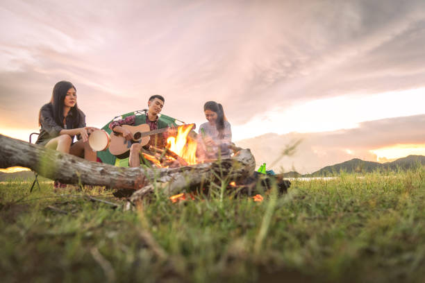 grupo de viajantes acampar e fazer piquenique e tocando juntos. fundo de montanha e lago. pessoas e estilo de vida. tema de atividade e lazer ao ar livre. mochileiros e alpinista. amanhecer e crepúsculo - bonfire people campfire men - fotografias e filmes do acervo