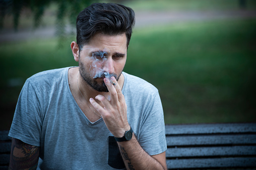 Depressed Young Adult Man sitting on a bench at public park smoking a cigarette