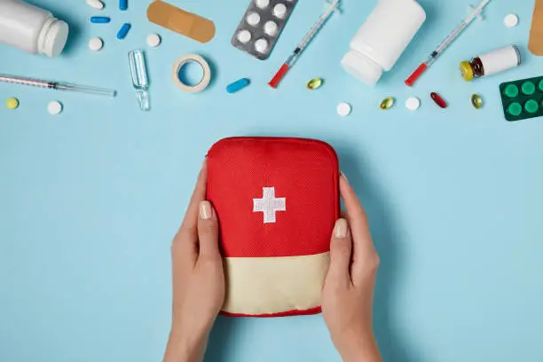 cropped shot of woman holding first aid kit bag over blue surface with various medicines