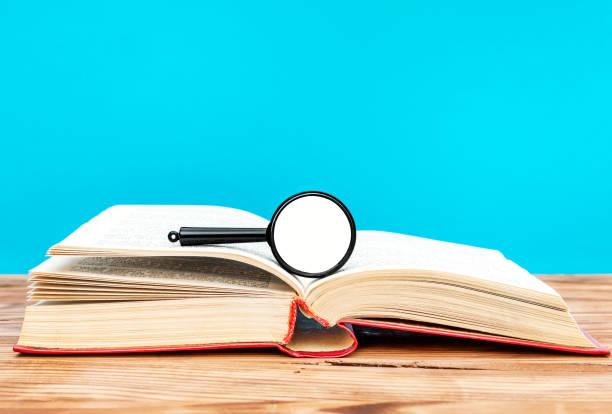 istock Opened book with magnification glass on wooden table against blue background. 992980582