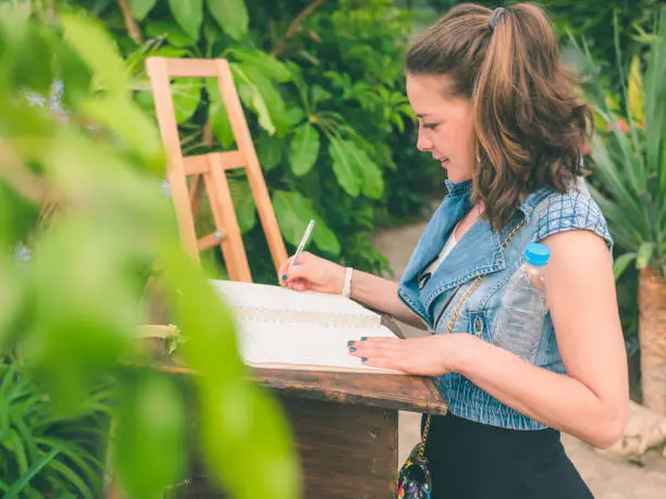 Cute girl writing in a guestbook in a beautiful garden. The young woman is of caucasian ethnicity and is casually dressed. The scene is situated in a botanical garden in Balchik, Bulgaria (Eastern Europe) around sunset. The photograph is take with Panasonic GH5 camera.