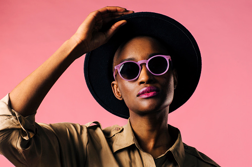 A fashion portrait of a young woman with purple sunglasses and black hat, isolated on pink studio background