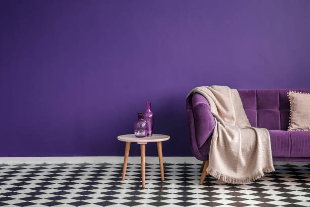 istock Dark purple sofa with a blanket beside a small table with bottles standing on black and white checkerboard floor in a minimalistic living room interior. Copy space. Real photo 993219808