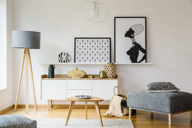 istock Wooden table and grey pouf in bright flat interior with posters and lamp next to cabinet. Real photo 993243224