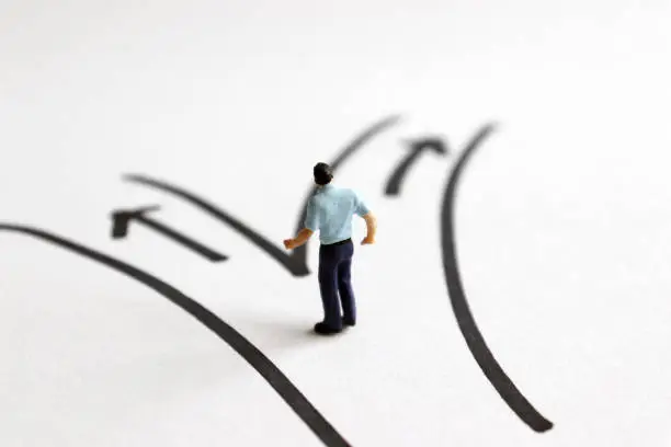 Photo of A miniature man standing in front of two different path.
