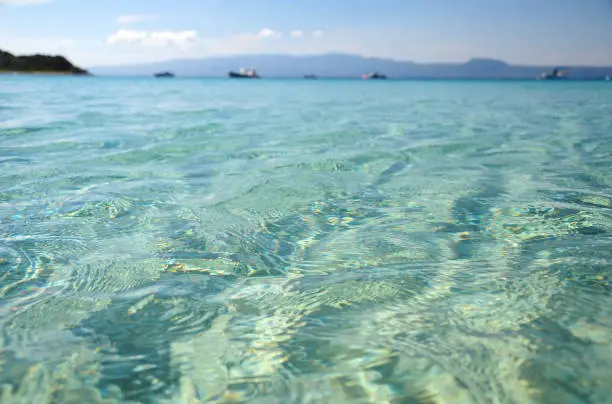 Transparent sea water surface close up with boats at background, Alikes beach, Ammouliani, Greece