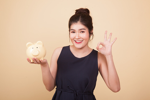 Asian woman show OK  with pig coin bank  on beige background