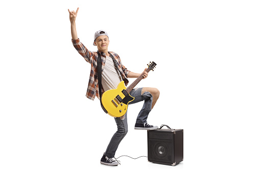 Full length portrait of a teenager with an electric guitar and an amplifier making a rock gesture isolated on white background