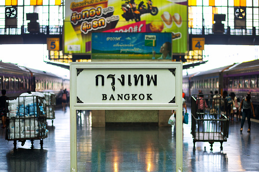 Bangkok station sign on platform in Hua Lamphong on center of platform with two tracks. At each side is a train. In background are some people and luggage carriers.