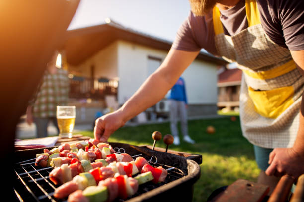 Dedicated man turning vegetables and meat on a stick to grill on other side as well while rest of the family is scattered around the house yard. Dedicated man turning vegetables and meat on a stick to grill on other side as well while rest of the family is scattered around the house yard. celebrity roast stock pictures, royalty-free photos & images