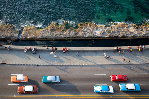 Large group of vintage American cars speeding along the Malecon in Havana, Cuba, motion blur, people sitting on sea wall, Caribbean Sea is visible in the background, elevated view.