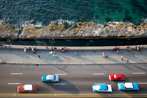 Group of vintage American cars speeding along the Malecon in Havana, Cuba, motion blur, people sitting on sea wall, Caribbean Sea is visible in the background, elevated view.