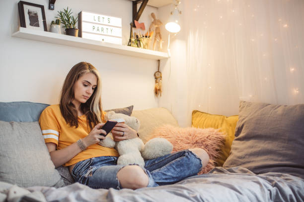 Weekend relaxation Young woman alone in bedroom. She work homework, using laptop, sitting on beed and holding teddy bear, drinking coffee, writing on papper and making selfie dorm room photos stock pictures, royalty-free photos & images