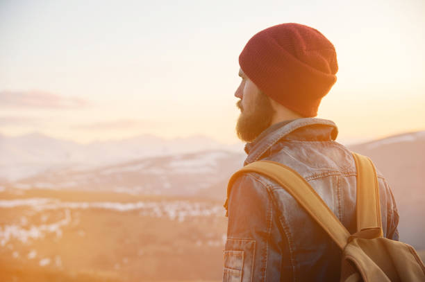 istock Bearded tourist hipster man in a hat with a backpack stand back on a roadside bump and watching the sunset against the background of a snow capped mountain 993781092