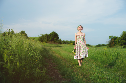 young happy smiling girl in a calico dress with a bouquet is walking along a country road with green grass.