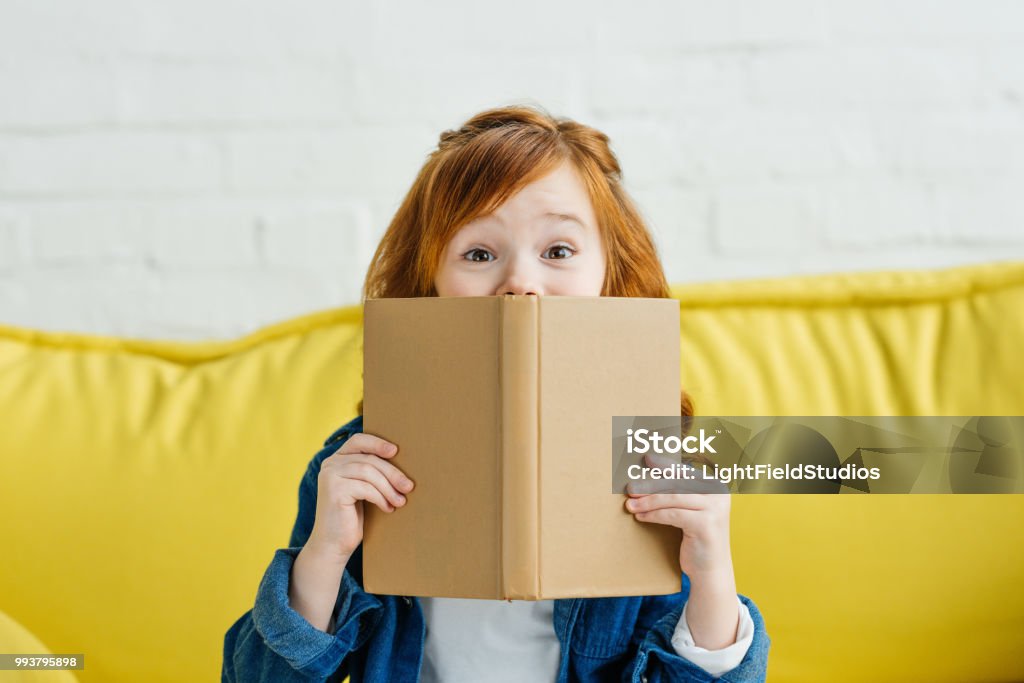 Child sitting on sofa and holding book in front of her face Child Stock Photo