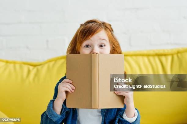 Child Sitting On Sofa And Holding Book In Front Of Her Face Stock Photo - Download Image Now