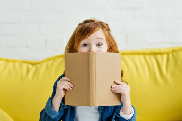 Photo of Child sitting on sofa and holding book in front of her face