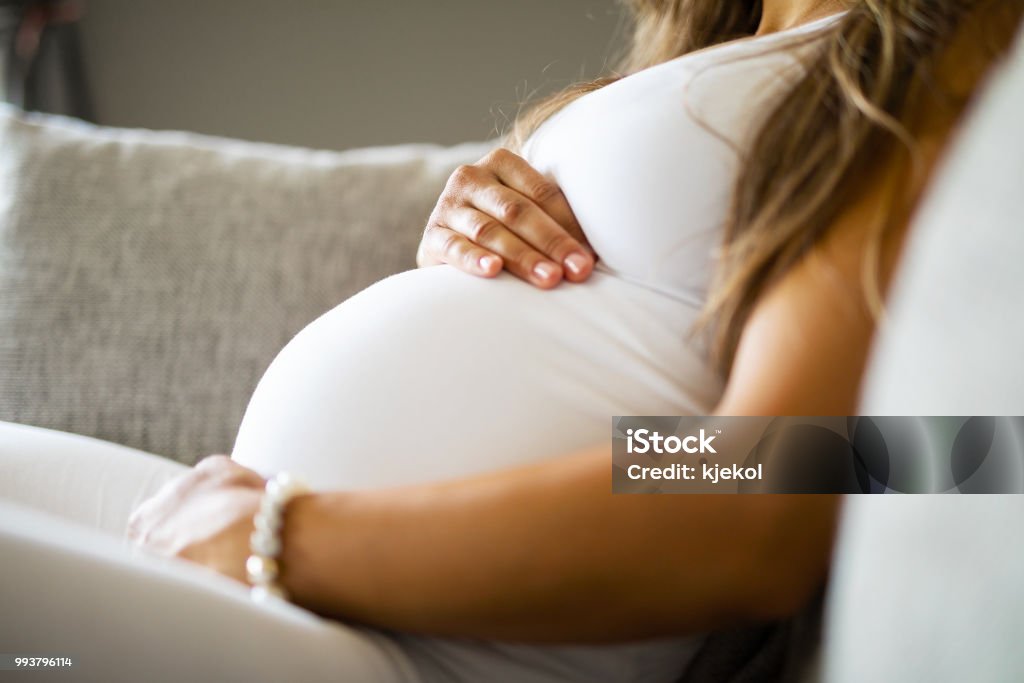 Close-up of pregnant woman sitting in sofa with her hands at belly Close-up of pregnant woman relaxing and sitting on the side on the sofa. Holding a hands on the tummy. Pregnant Stock Photo