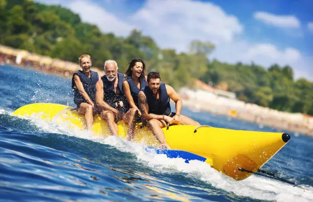 Closeup of senior couple with their children enjoying banana boat ride during their summer vacation. They are excited and hardly holding onto the handles, but it's big fun, especially for the parents.