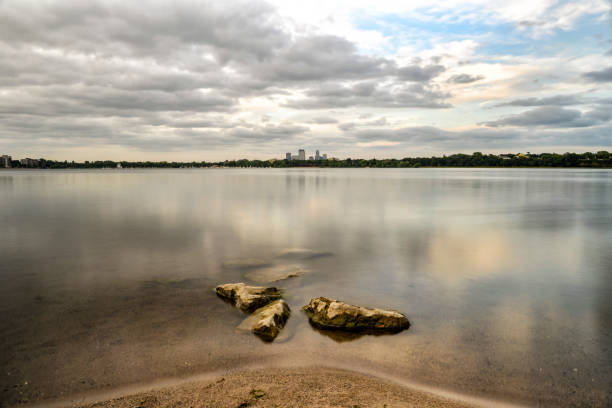 Lake, Beach and Rocks with Minneapolis Skyline in Background with Calm Lake Water and Cloudy Sky.