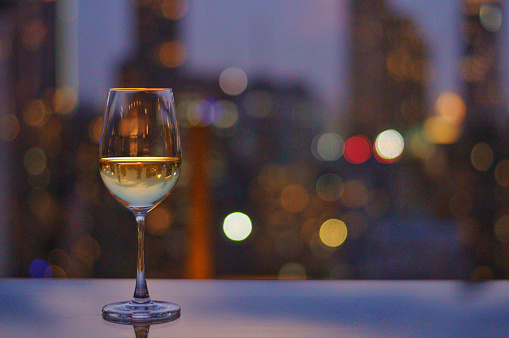 A glass of white wine on table of rooftop bar with colorful bokeh of city light and space for text.