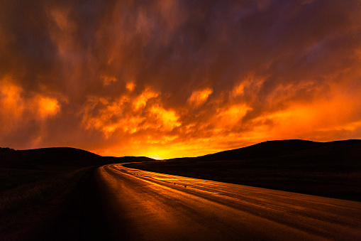 Wet fiery red highway reflecting storm clouds at sunset curving in South Dakota hills